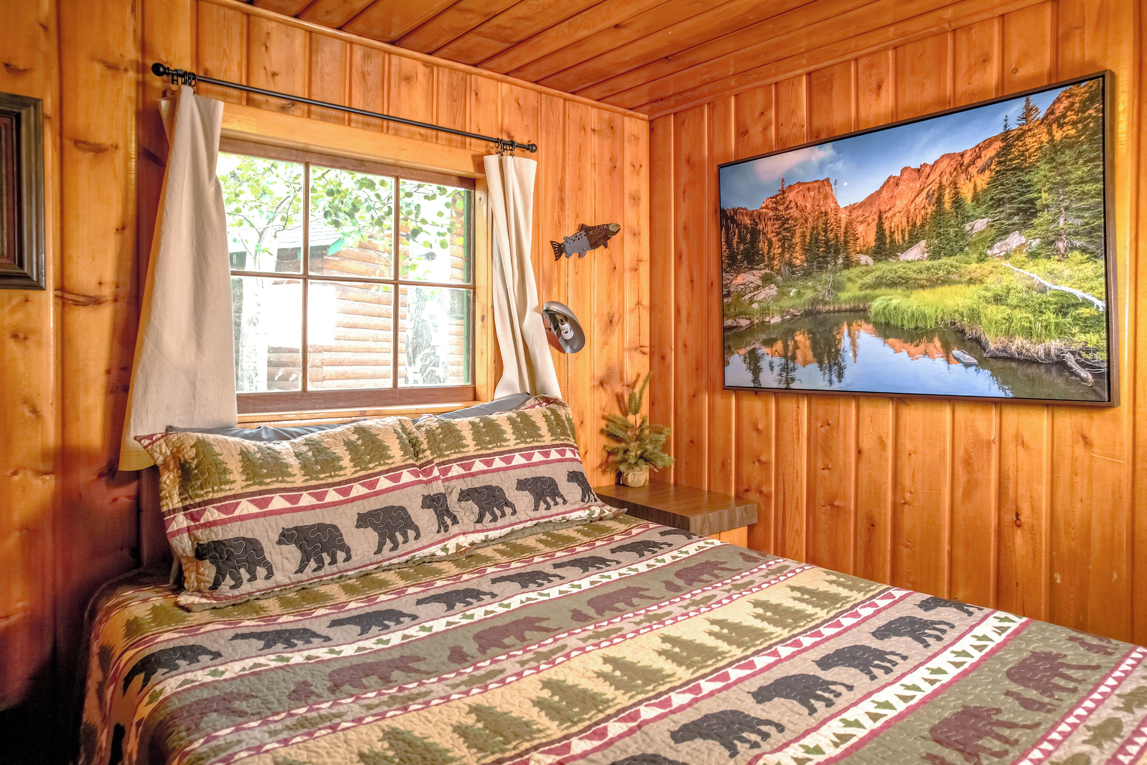 Una habitación de cabaña de madera con una cama, una ventana con cortinas, un adorno de pez en la pared y un cuadro de un paisaje montañoso.