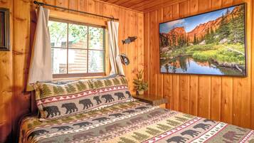 Una habitación de cabaña de madera con una cama, una ventana con cortinas, un adorno de pez en la pared y un cuadro de un paisaje montañoso.