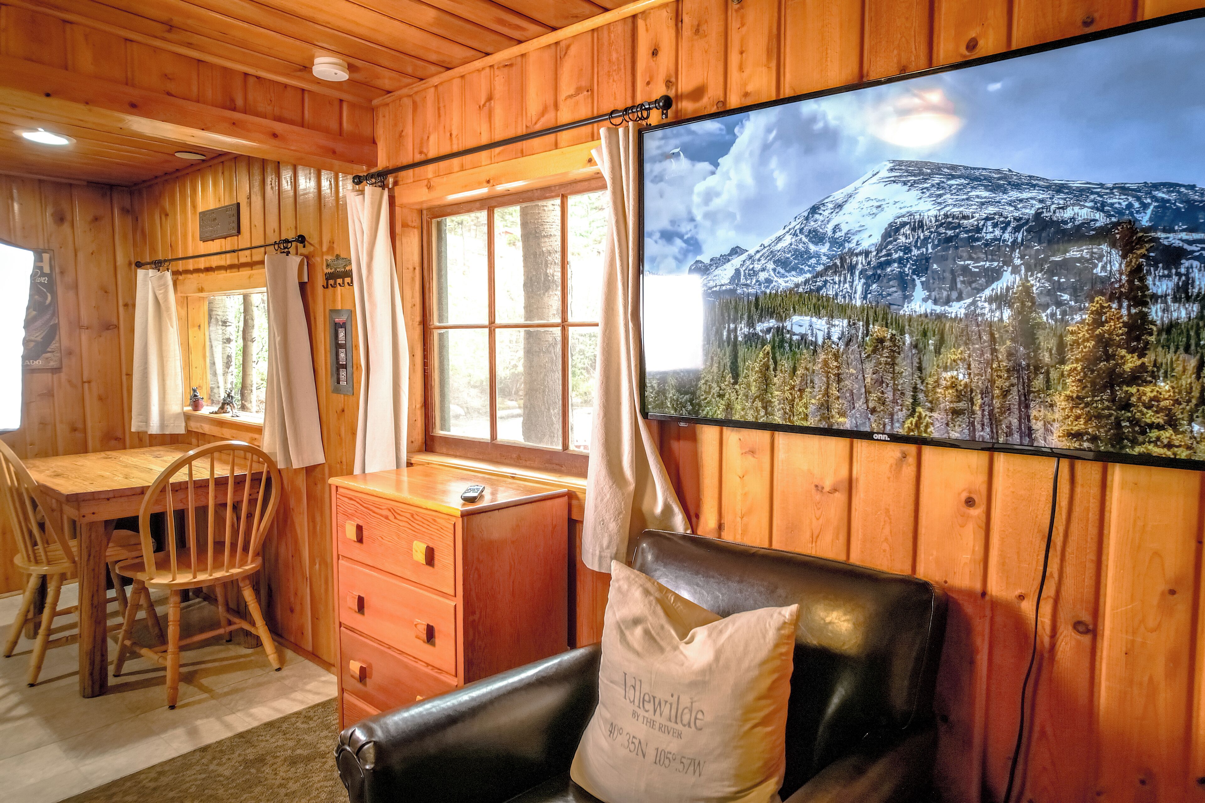 A cabin interior with a wooden interior, a dining table with chairs, a desk with a chair, a window with curtains, and a large framed picture of a mountain landscape.
