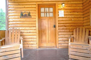 L'ingresso di una baita di legno, con una porta, un cartello di benvenuto e due panche in legno su entrambi i lati.