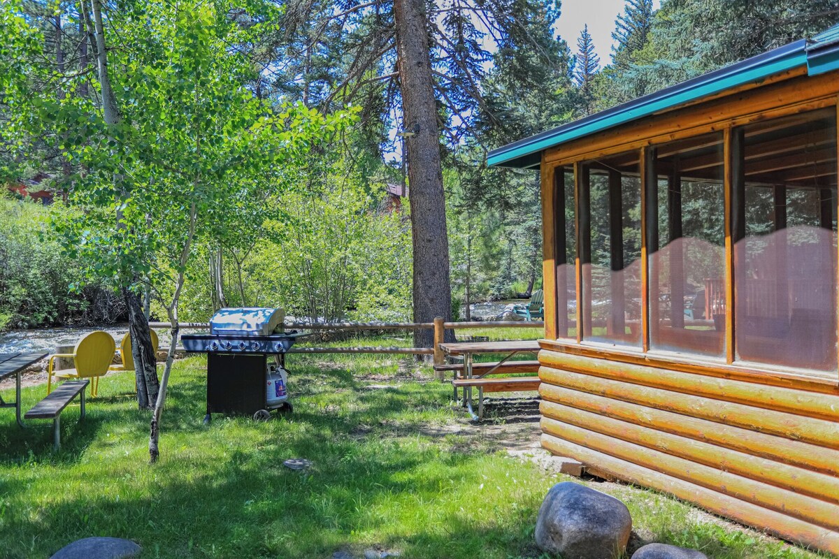 A rustic cabin with a wooden exterior and large windows, surrounded by a grassy area with picnic tables and a barbecue grill.