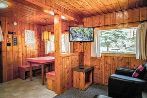 Interior de una cabaña con un televisor montado en la pared, una mesa de madera, un banco y una ventana con cortinas.