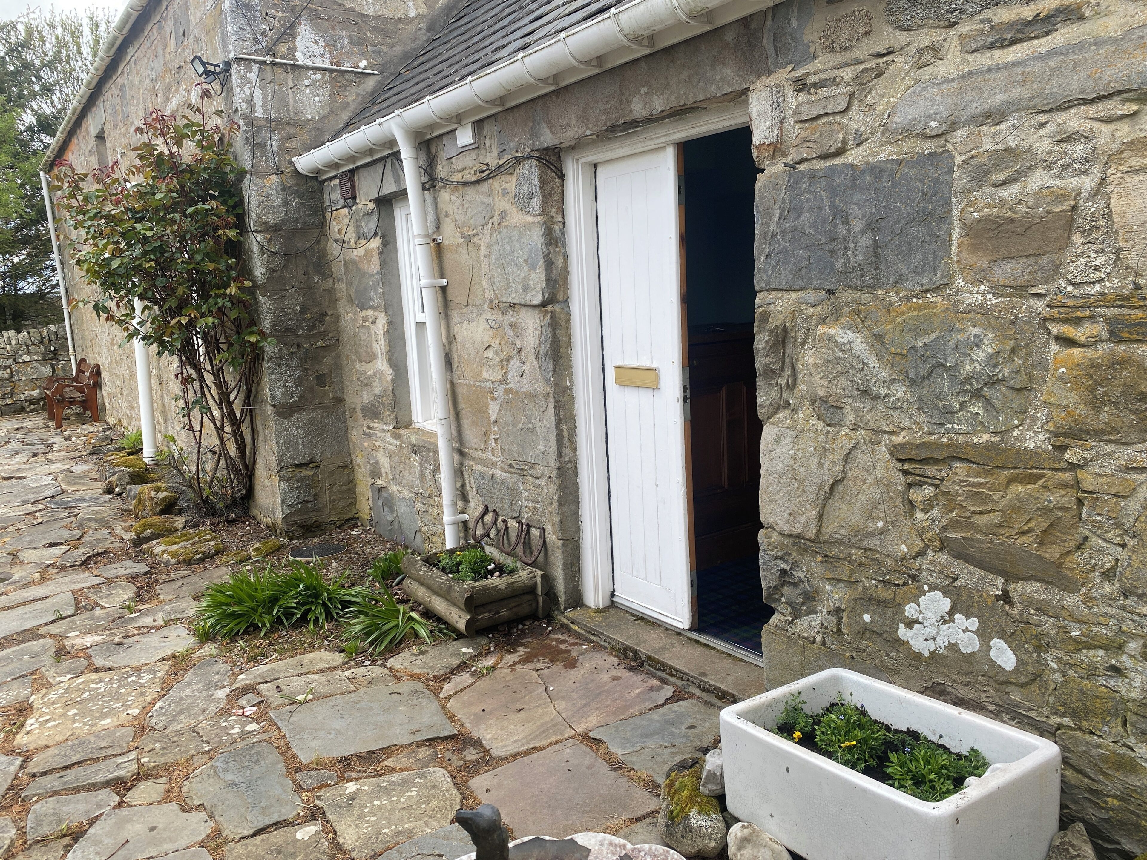 Un edificio de piedra con una puerta blanca, un pequeño jardín con plantas y un camino de piedra.