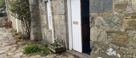 Un edificio de piedra con una puerta blanca, un pequeño jardín con plantas y un camino de piedra.