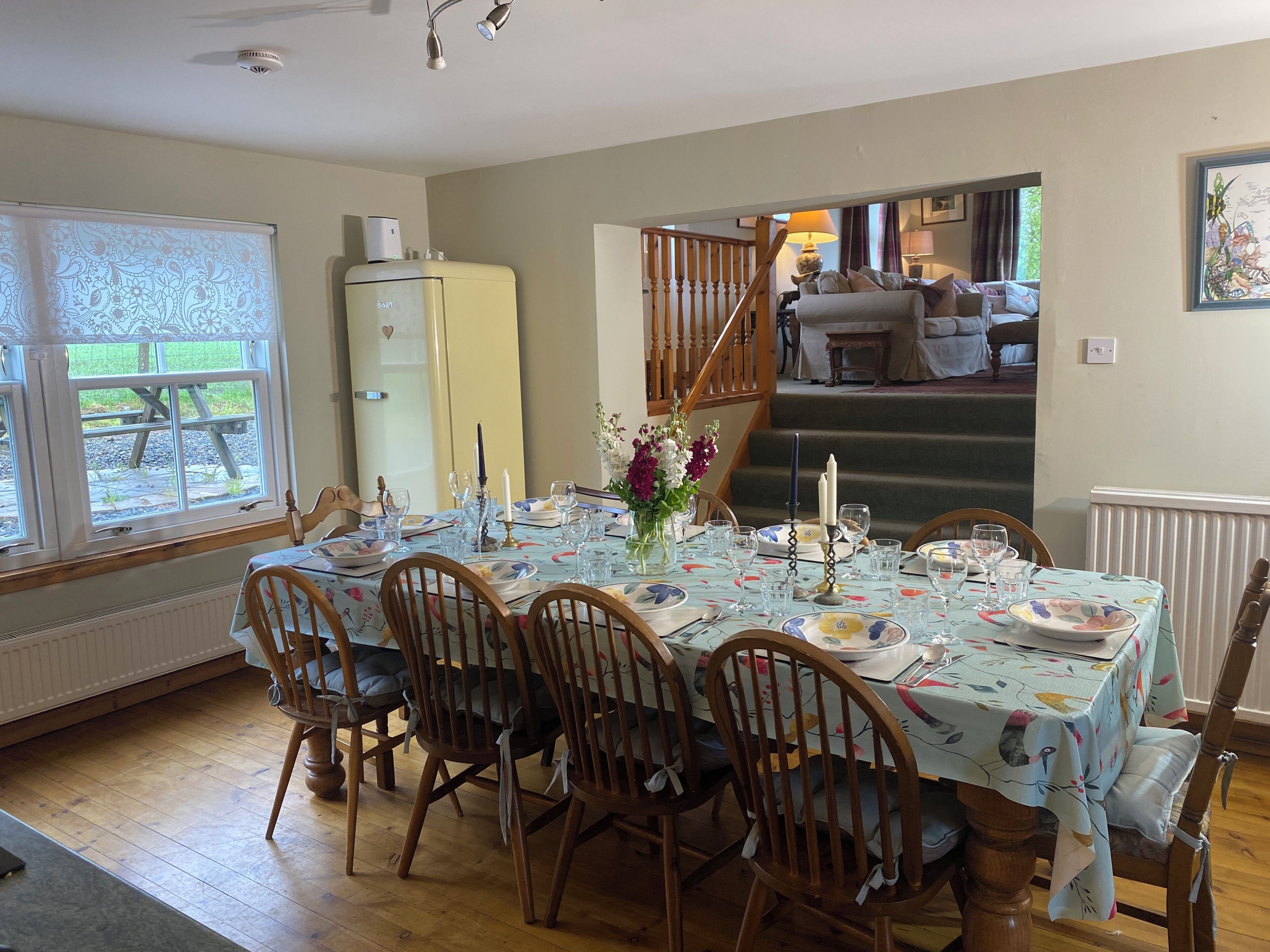 A dining room with a table set for six, wooden chairs, a refrigerator, a staircase, and a view of the outdoors.