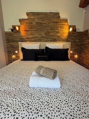 Un lit avec une couvre-lit à motifs, deux oreillers, une serviette enroulée et une tête de lit en bois agrémentée de lampes fixées au mur.