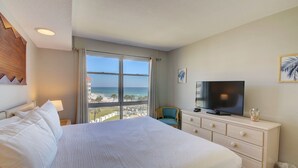 Een hotelkamer met een groot bed, een televisie, een dressoir en uitzicht op het strand.
