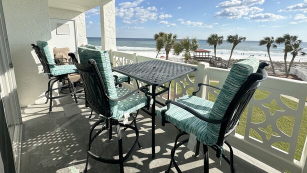 A balcony with a table and chairs, overlooking a beach with palm trees and a pier.
