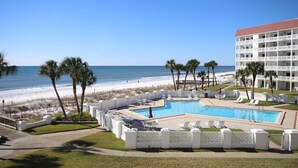 A pool area with a view of the beach and ocean, surrounded by palm trees and a white fence.