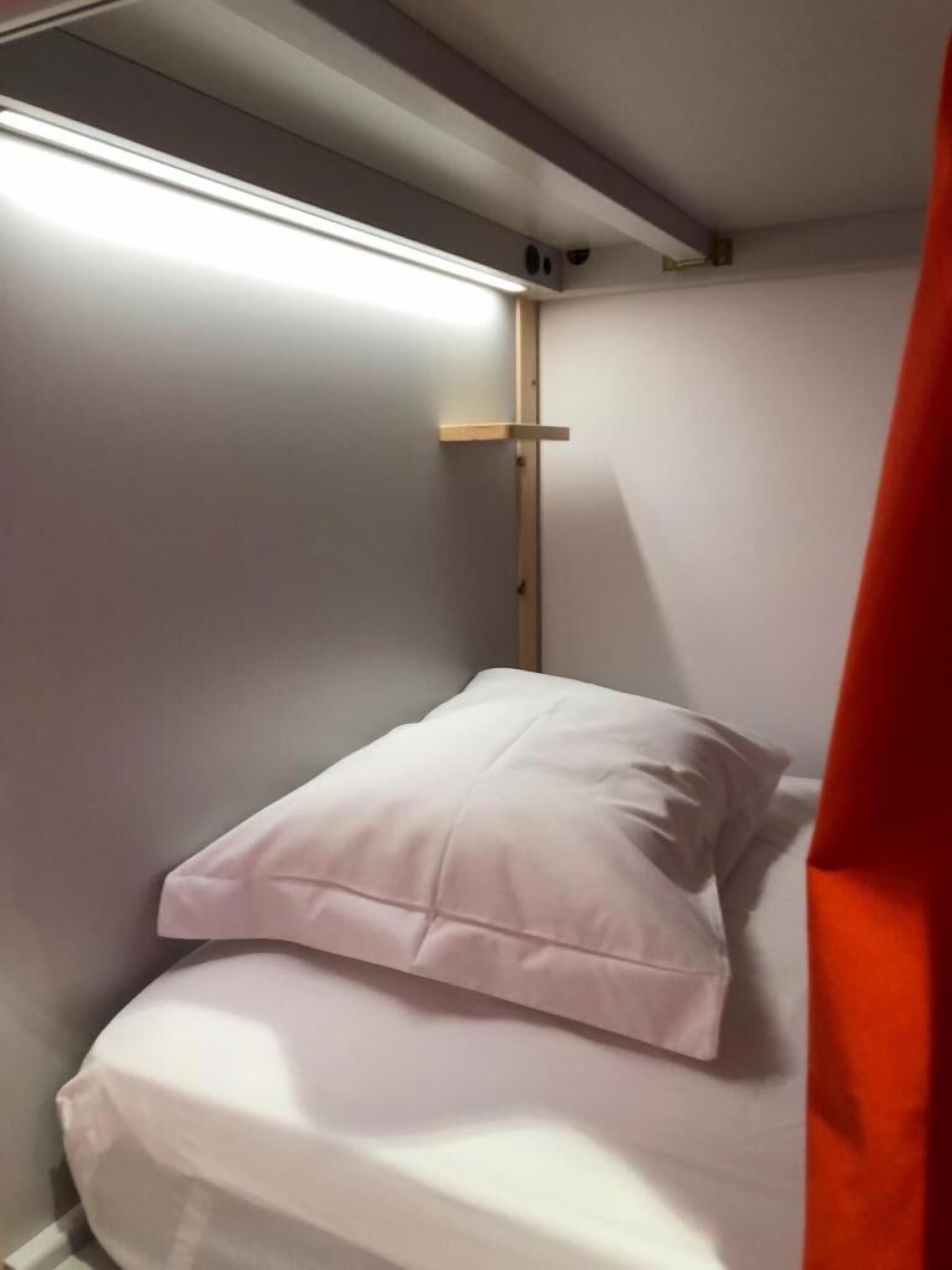 A small, narrow room with a single bed, a wooden shelf, and a red curtain.