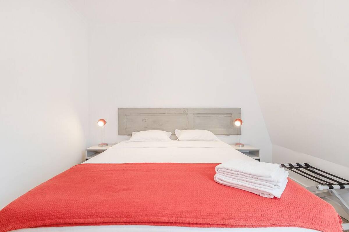 Una cama bien tendida con una manta roja, almohadas blancas y una toalla blanca doblada al lado. Hay dos lámparas de noche con pantallas rojas y un cabecero de madera.