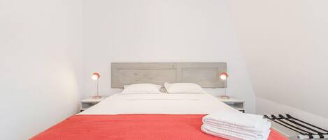 Una cama bien tendida con una manta roja, almohadas blancas y una toalla blanca doblada al lado. Hay dos lámparas de noche con pantallas rojas y un cabecero de madera.
