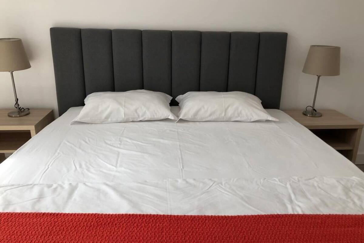 Una cama con ropa blanca, un cabecero tapizado en gris y dos lámparas de noche sobre mesitas de madera.
