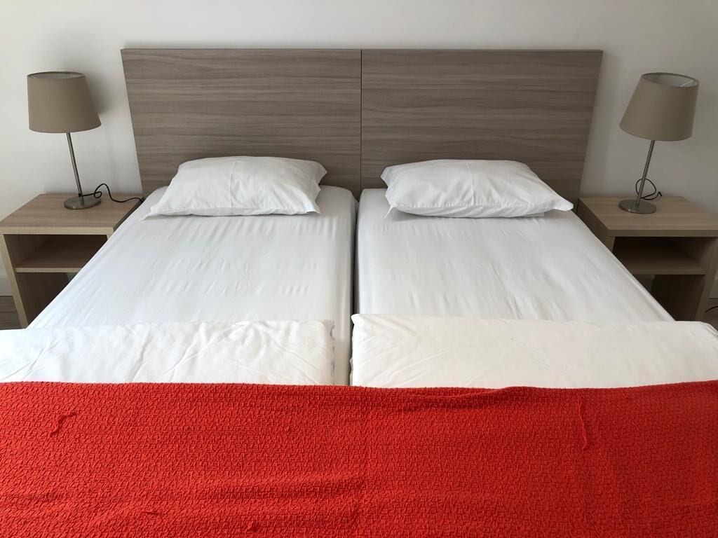 Una cama doble con sábanas blancas y una manta roja, dos mesitas de noche con lámparas, y un cabecero de madera.