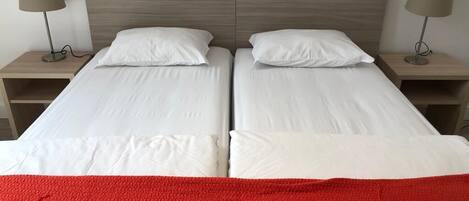 Una cama doble con sábanas blancas y una manta roja, dos mesitas de noche con lámparas, y un cabecero de madera.