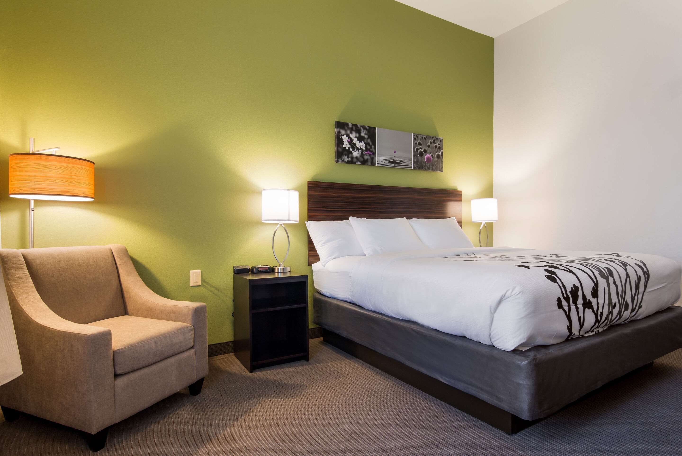 Una habitación de hotel con una cama, una silla, dos mesitas de noche con lámparas y una pared con un cuadro.