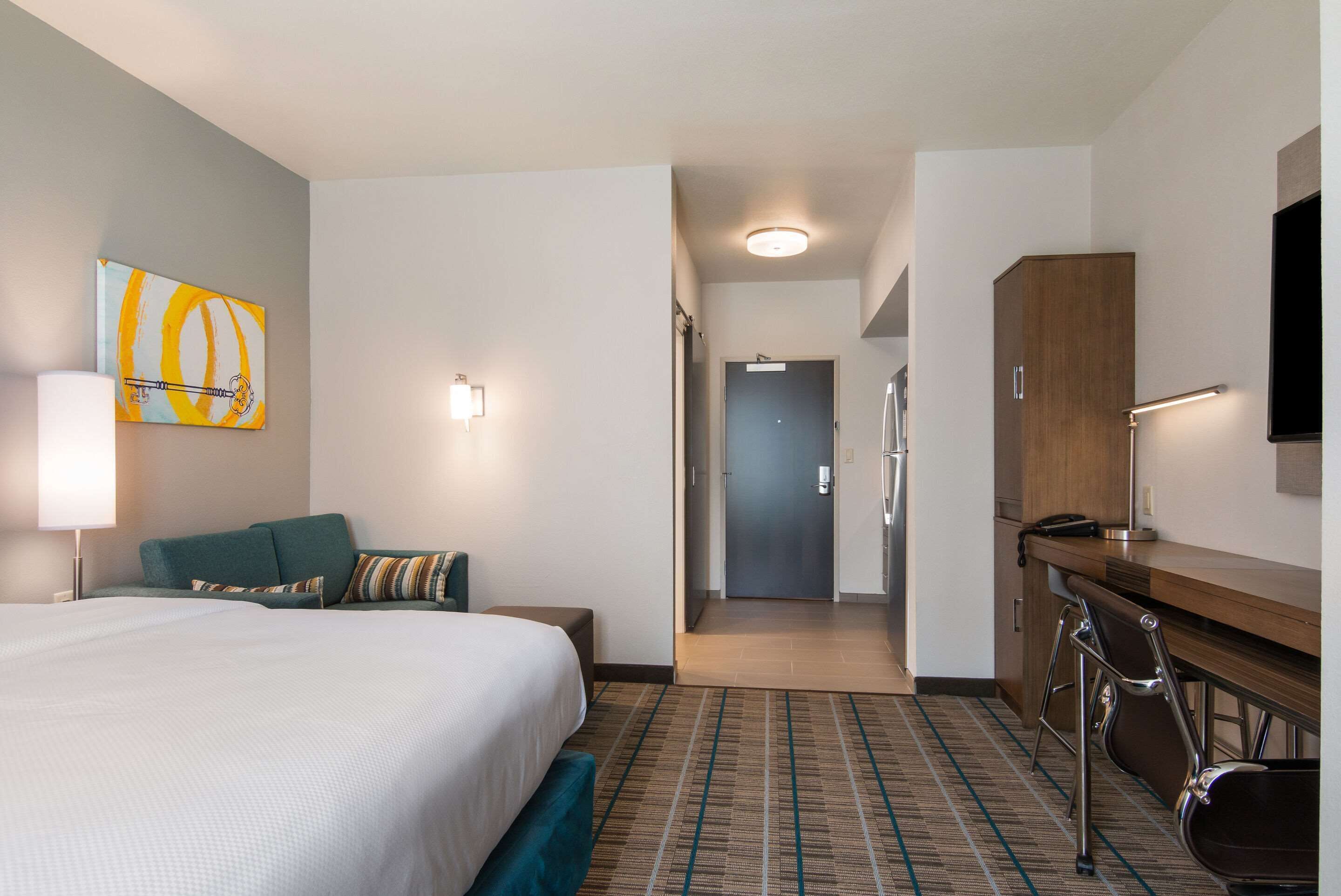 A hotel room with a bed, a desk, a chair, a television, and a door leading to a hallway.
