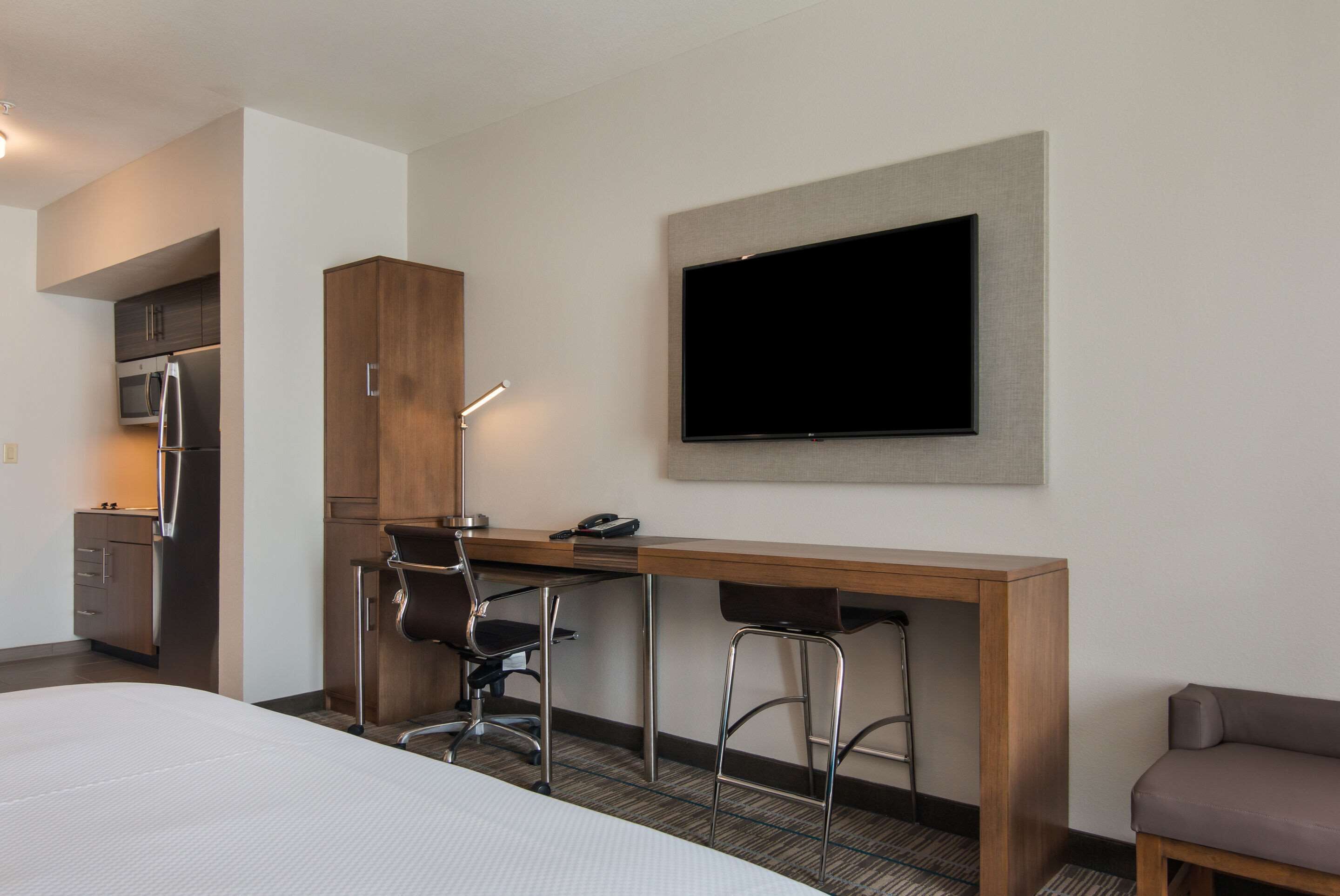 Habitación de hotel con cama, escritorio con dos sillas, televisor de pantalla plana montado en la pared, kitchenette con microondas y refrigerador, además de un sofá gris.