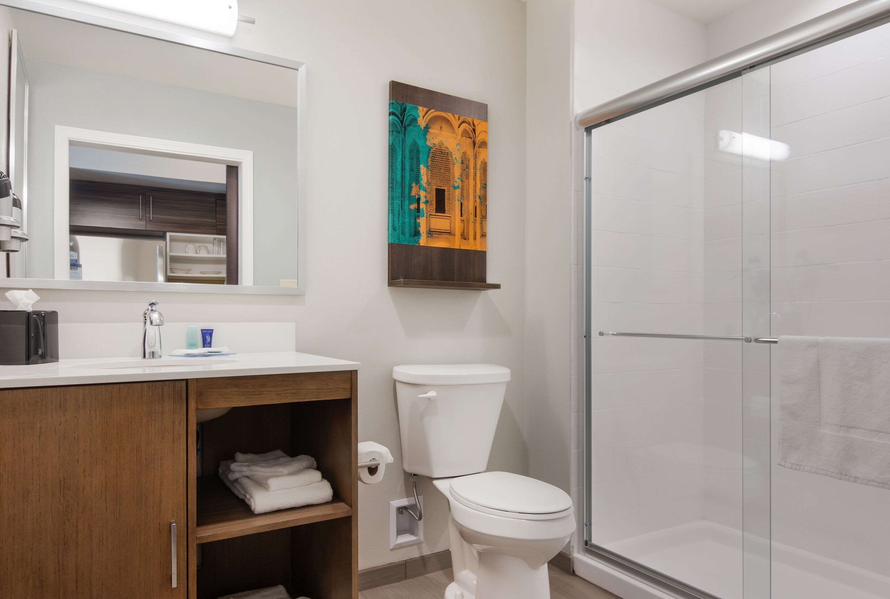 Un baño con inodoro blanco, cabina de ducha de vidrio, tocador de madera con espejo y una obra de arte en la pared.