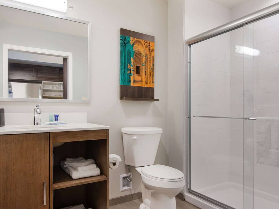 Un baño con inodoro blanco, cabina de ducha de vidrio, tocador de madera con espejo y una obra de arte en la pared.