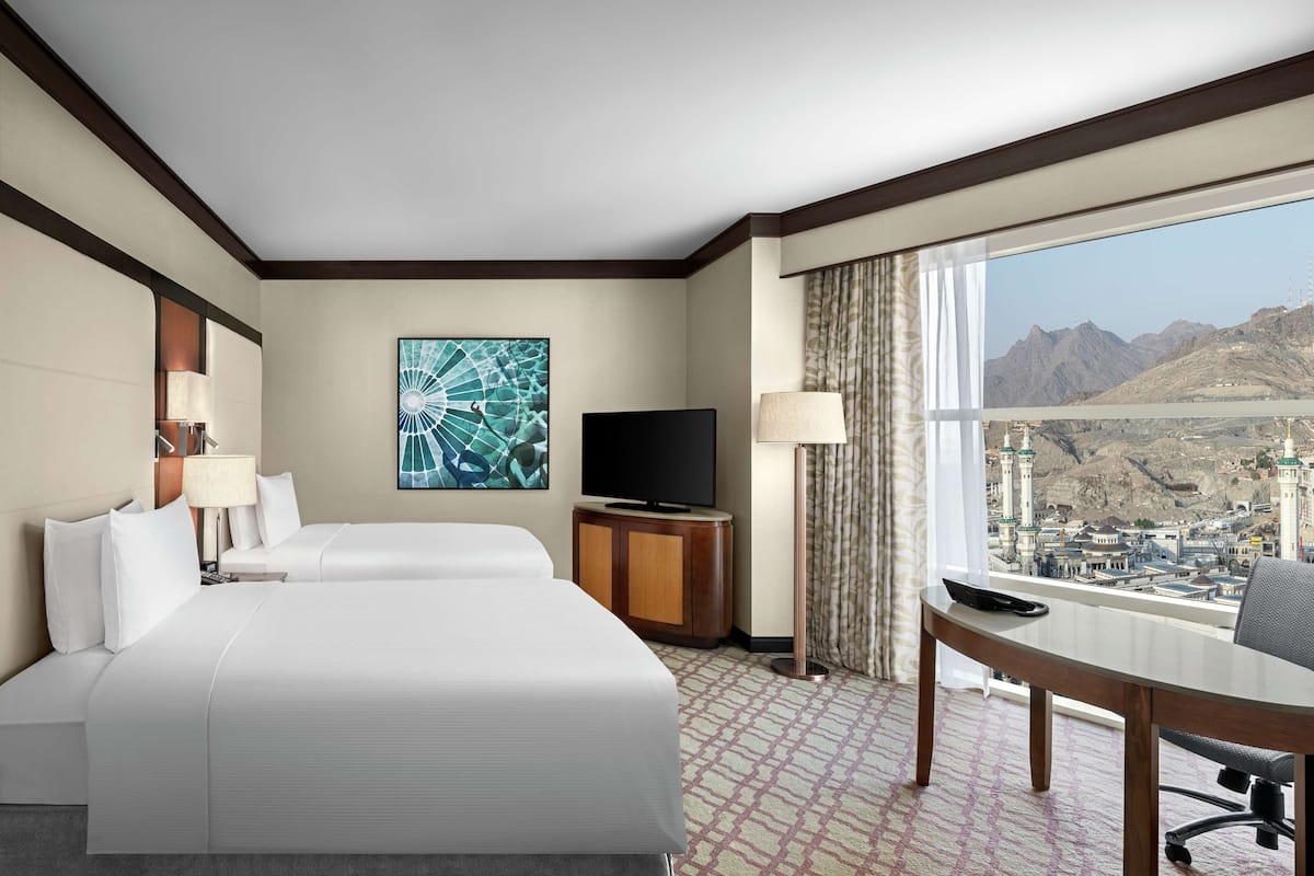 A hotel room with a large bed, a TV, a desk with a chair, and a view of a cityscape.
