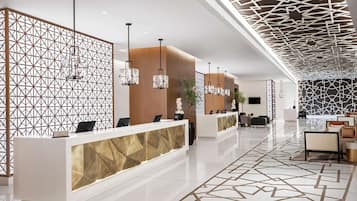 Un hall d’hôtel moderne avec un mur orné d’un motif géométrique, un comptoir d’accueil agrémenté d’une touche dorée et des luminaires suspendus.