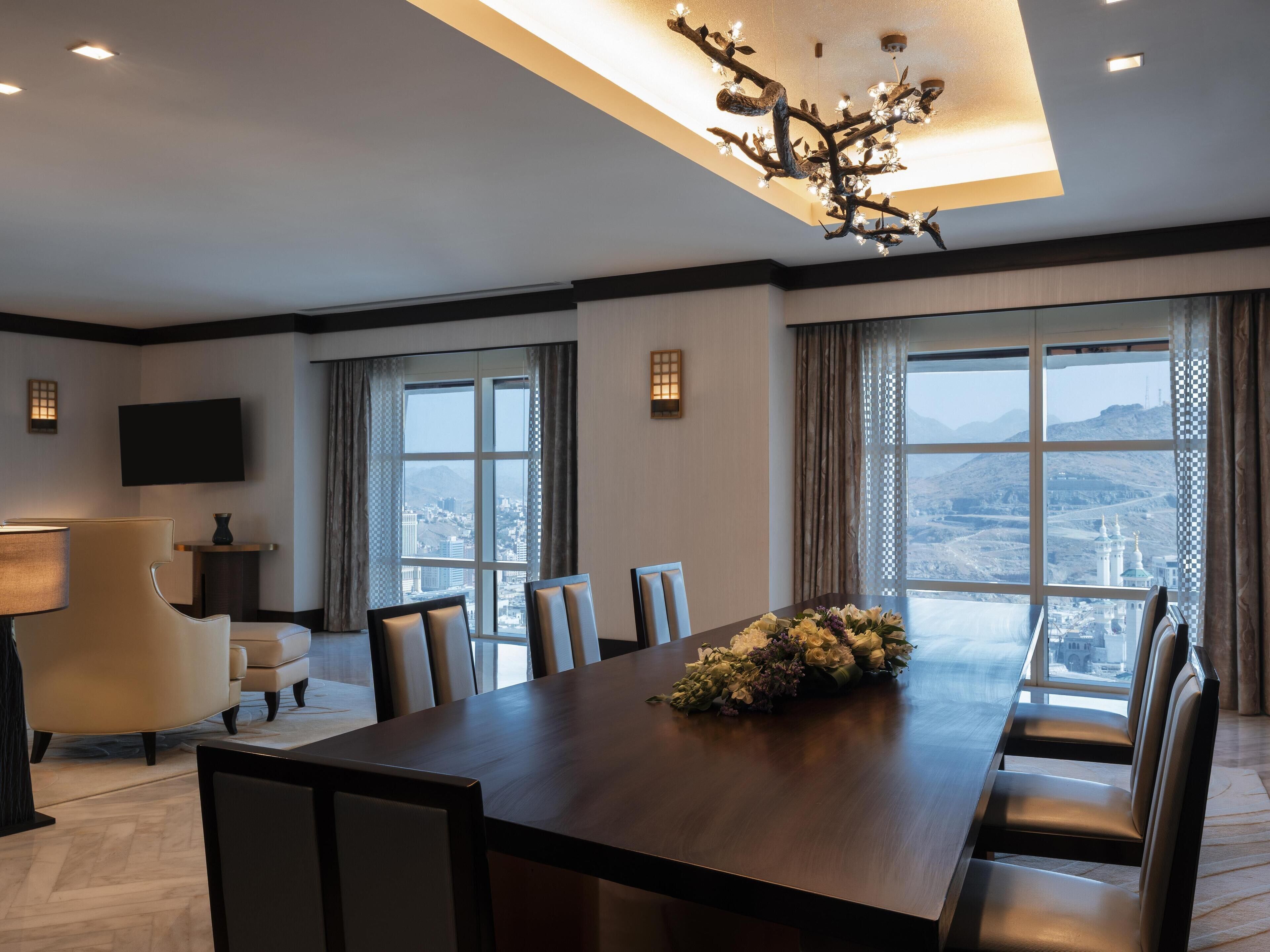 A modern hotel room with a large dining table, comfortable seating, a flat-screen TV, and large windows offering a city view.