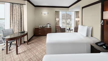 A hotel room with a large bed, a desk, a chair, a small table with a lamp, and a view of the city.