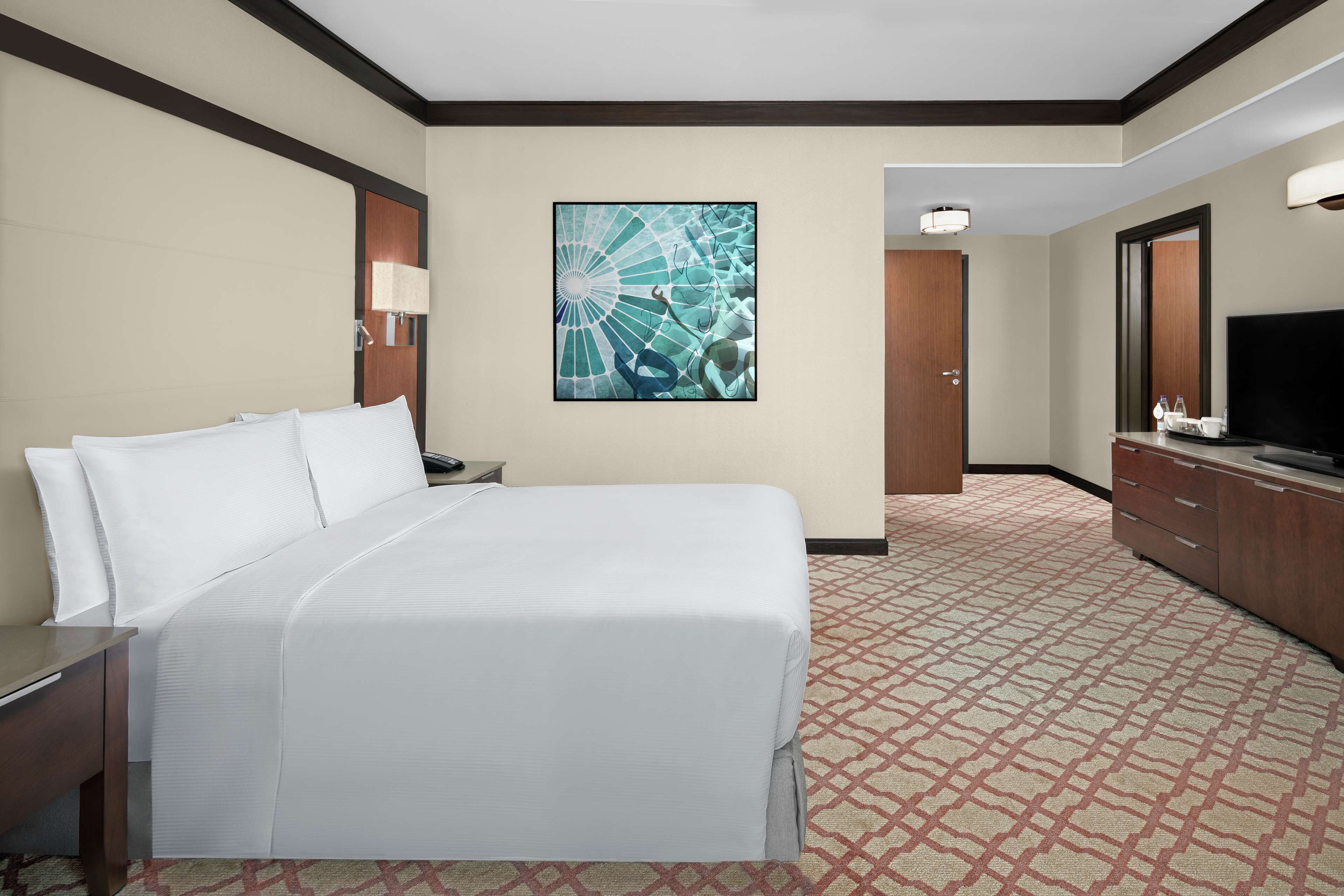 Ein Hotelzimmer mit einem großen Bett, einem Schreibtisch, einem Fernseher, einem Spiegel und einem Wandbild.