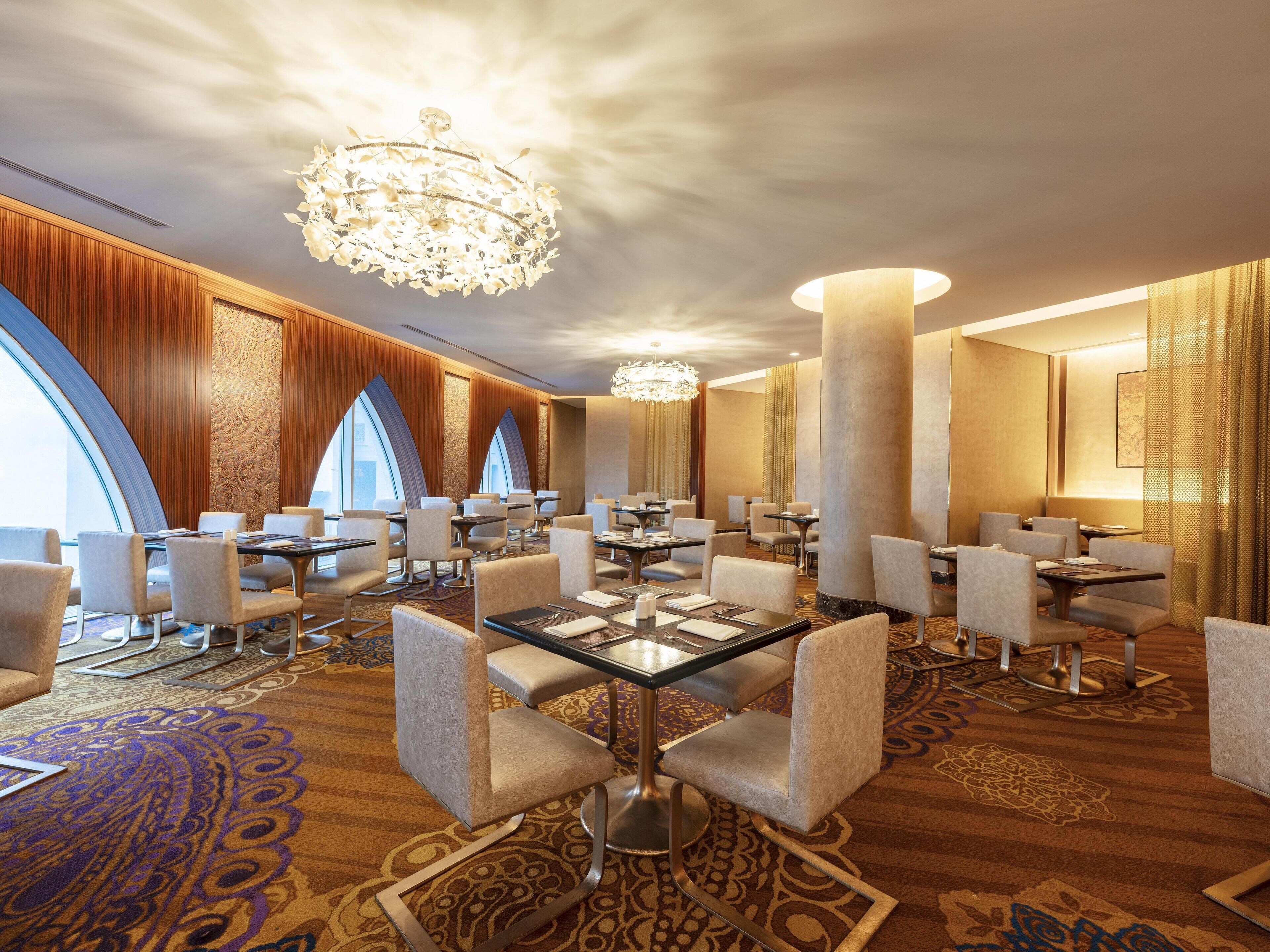 A hotel dining area with round tables, comfortable chairs, a large chandelier, and a view of the city through large windows.