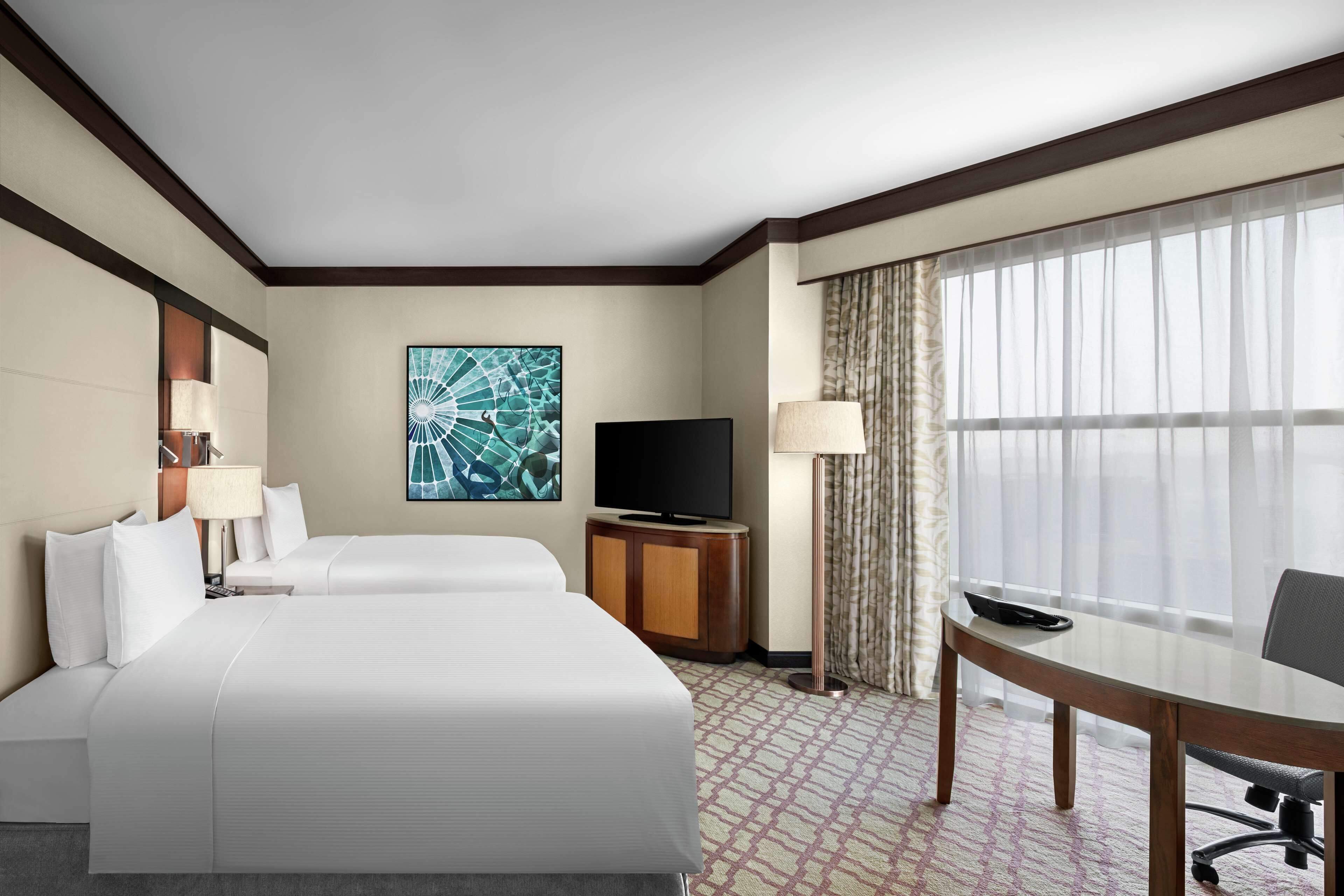 A hotel room with a large bed, a TV, a desk with a chair, and a window with sheer curtains.