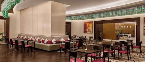 A spacious dining area with multiple tables and chairs, a central seating area with sofas and red cushions, and a patterned carpet.