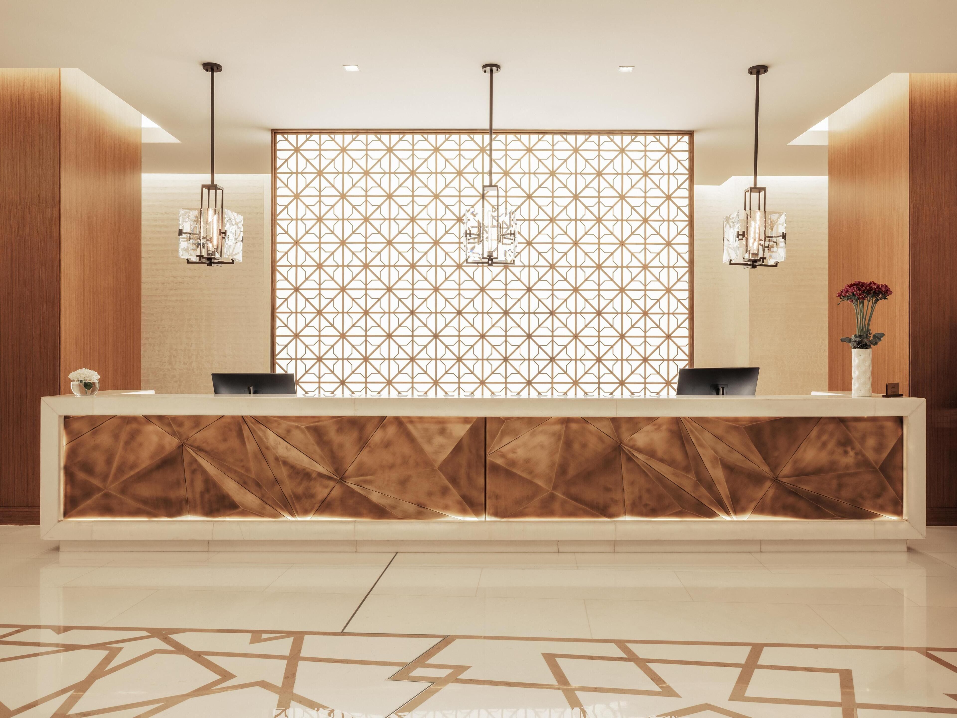 A modern reception desk with geometric patterned backdrop, two pendant lights, and a vase with flowers.