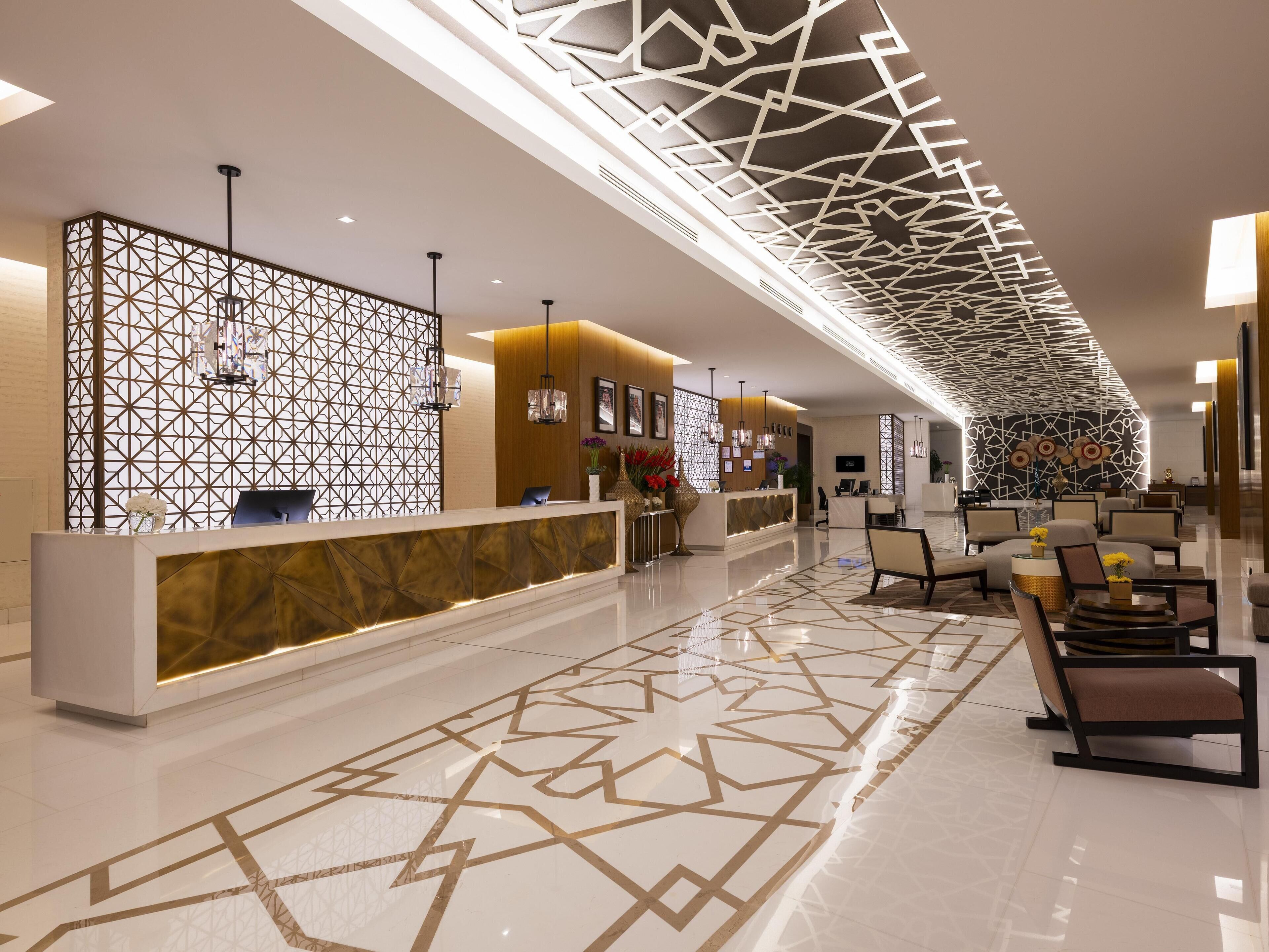 Un moderno vestíbulo de hotel con un mostrador de recepción de diseño geométrico, zona de estar y un plafón con lámpara geométrica.