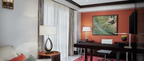 Habitación de hotel con sofá, escritorio, silla, lámpara, un cuadro y ventana con cortinas.