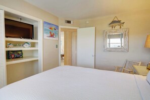 Een slaapkamer met een bed, een televisie op een plank, een stoel, een bureau en een deur naar een andere kamer.