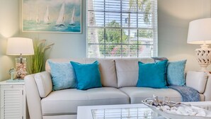 Ein Wohnzimmer mit einem beigen Sofa, türkisfarbenen Kissen, einem Glastisch und einem Bild von Segelbooten.