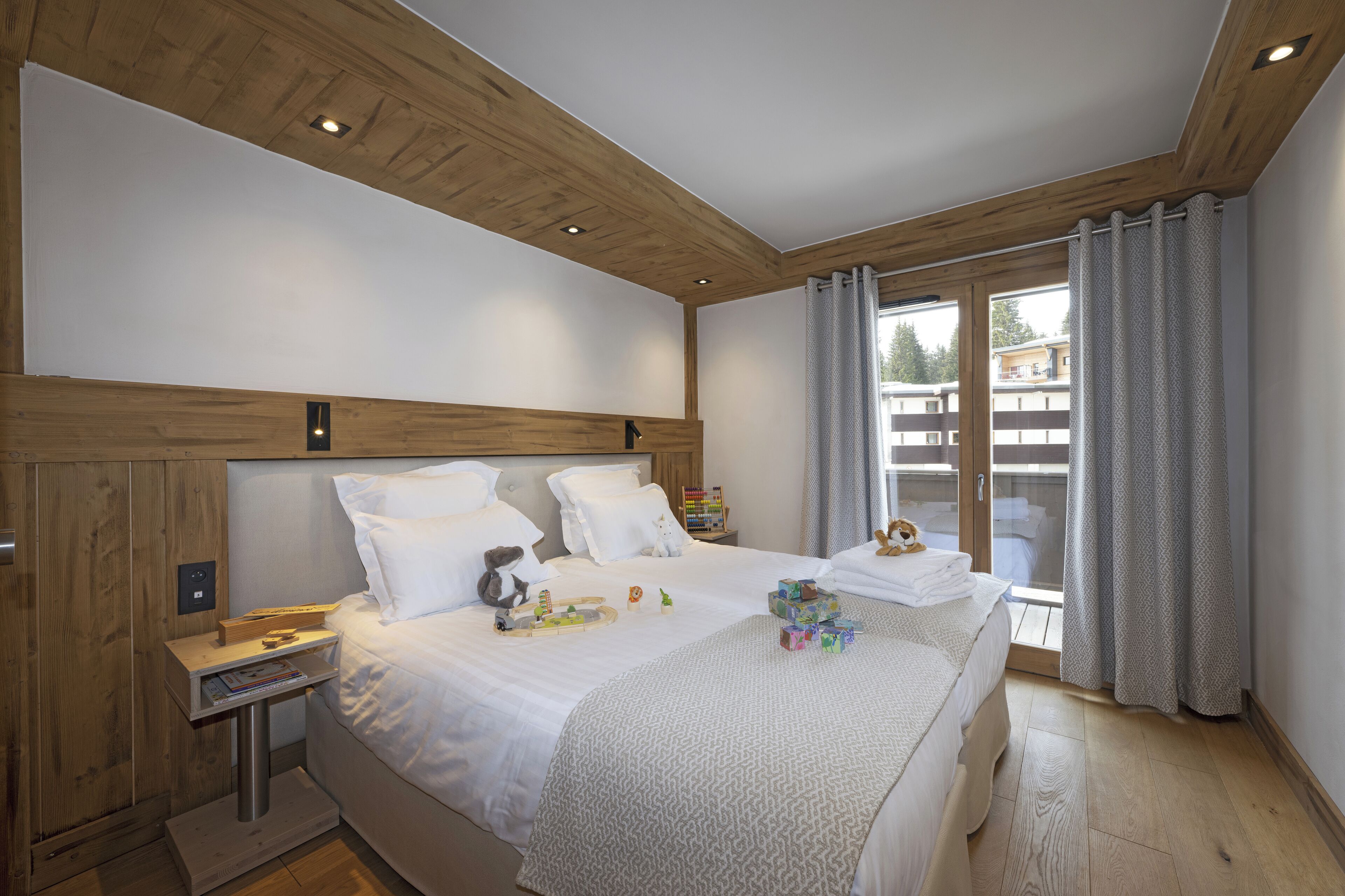 Une chambre moderne avec un plafond en bois, un grand lit avec une literie blanche, une table de chevet avec une lampe, et une vue sur un immeuble à travers une porte vitrée.