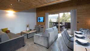 A modern living room with a sofa set, a coffee table, and a TV mounted on the wall. - MGM Hôtels & Résidences – Résidence Amaya (Villard-sur-Doron)