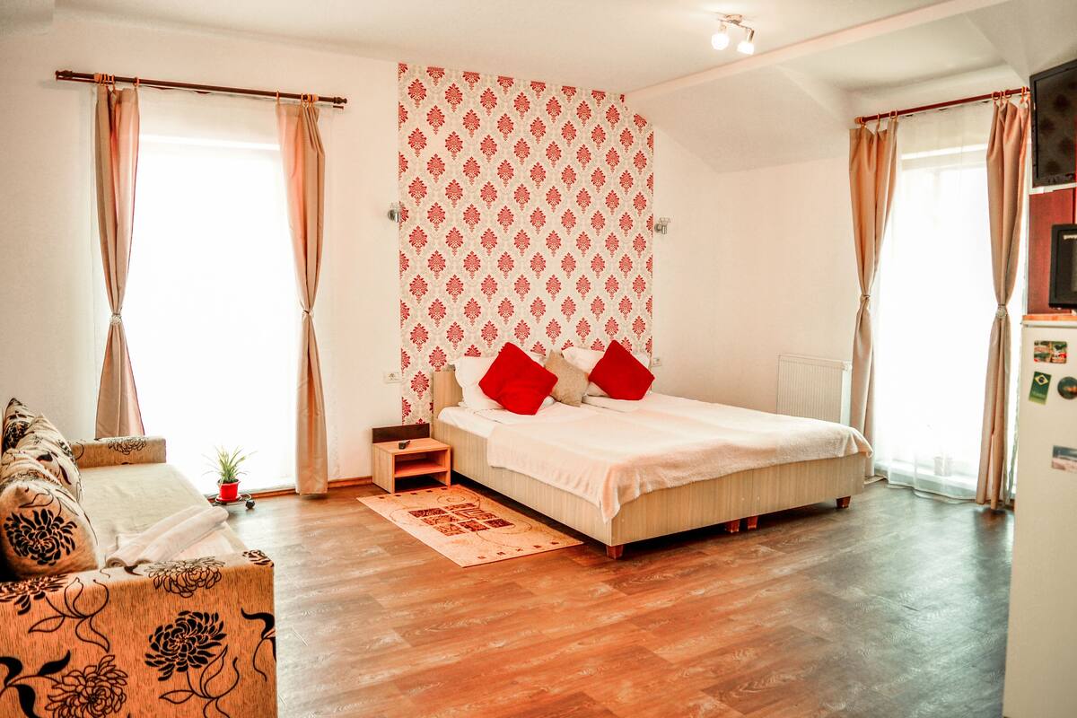 Ein Schlafzimmer mit Bett, Vorhängen, einem kleinen Tisch, einer Pflanze und Holzboden.