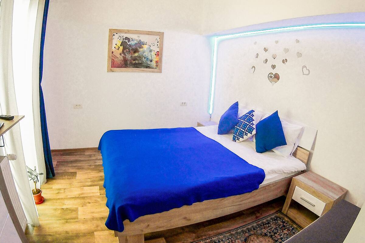 Ein Schlafzimmer mit einem hölzernen Bett, blauer Bettwäsche, einem kleinen Nachttisch, einer Wandleuchte und einem Bild an der Wand.