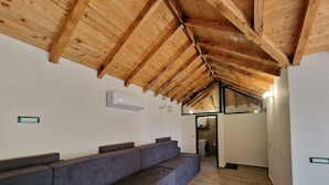 Une pièce avec un plafond en bois, un canapé gris, un climatiseur fixé au mur et une porte donnant sur une autre pièce.