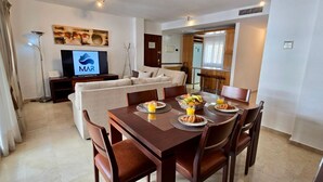 A modern hotel room with a dining area featuring a wooden table set for breakfast, a sofa, and a television displaying the MAR logo. - Urban 2 BR apartment : Pool : Los Boliches (Fuengirola)