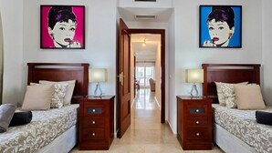 A hotel room with two beds, each with a wooden headboard, a bedside table, and a lamp. There are two framed artworks on the walls, a door leading to another room, and a hallway visible in the background. - Urban 2 BR apartment : Pool : Los Boliches (Fuengirola)