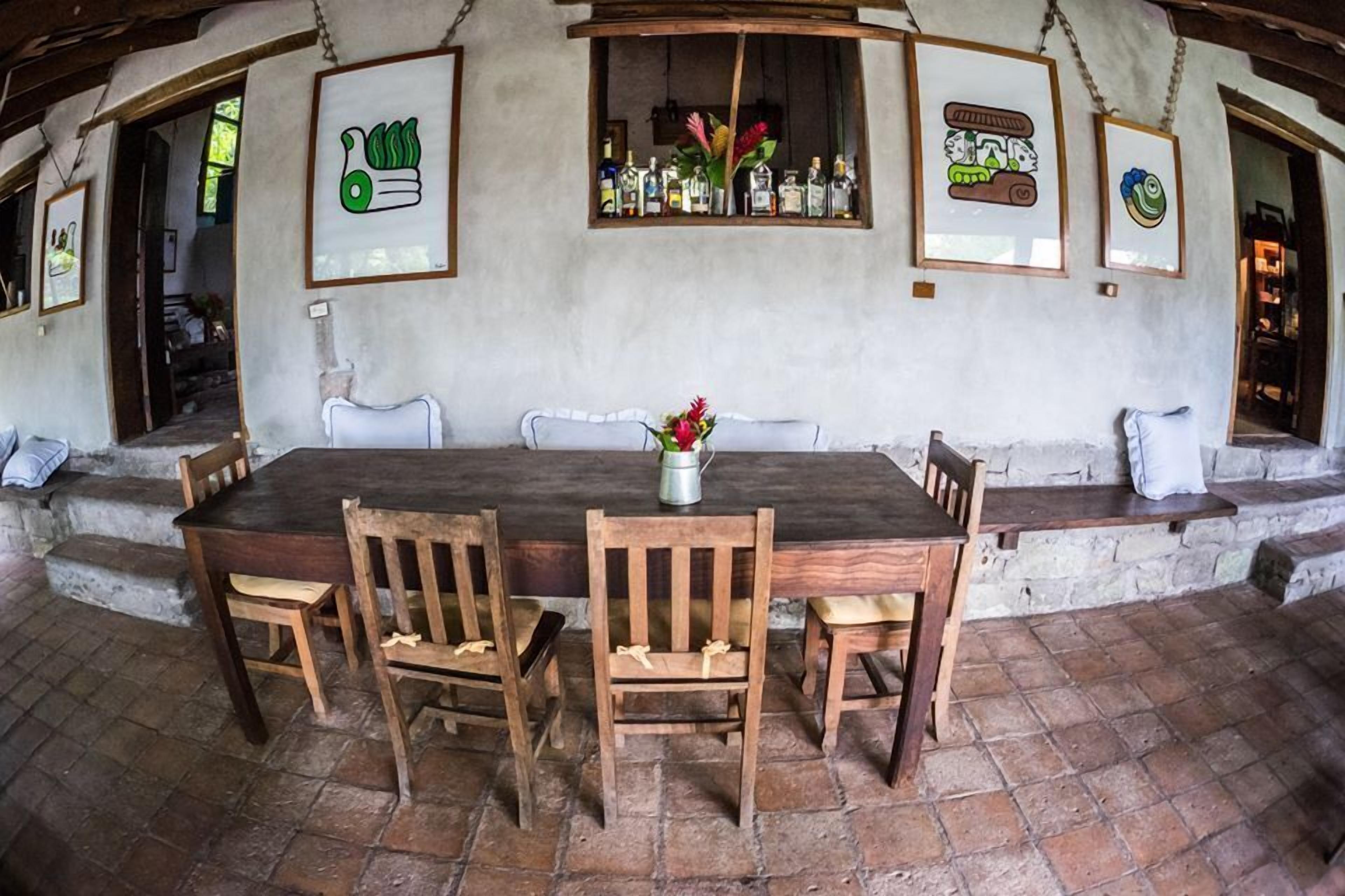 A rustic dining area with a wooden table and chairs, a stone floor, and framed artwork on the walls.