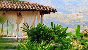 Un bâtiment traditionnel avec un toit en tuiles et un porche abrité, entouré d’une végétation luxuriante et avec des montagnes en arrière-plan.