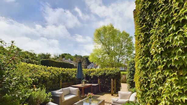 Eine Terrassengruppe mit Korbmöbeln, einem Glastisch und Blick auf einen Garten mit Hecke und Baum.