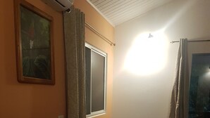 Une pièce avec une table en bois, un canapé gris, une chaise en osier, une fenêtre avec des rideaux et un luminaire au plafond.