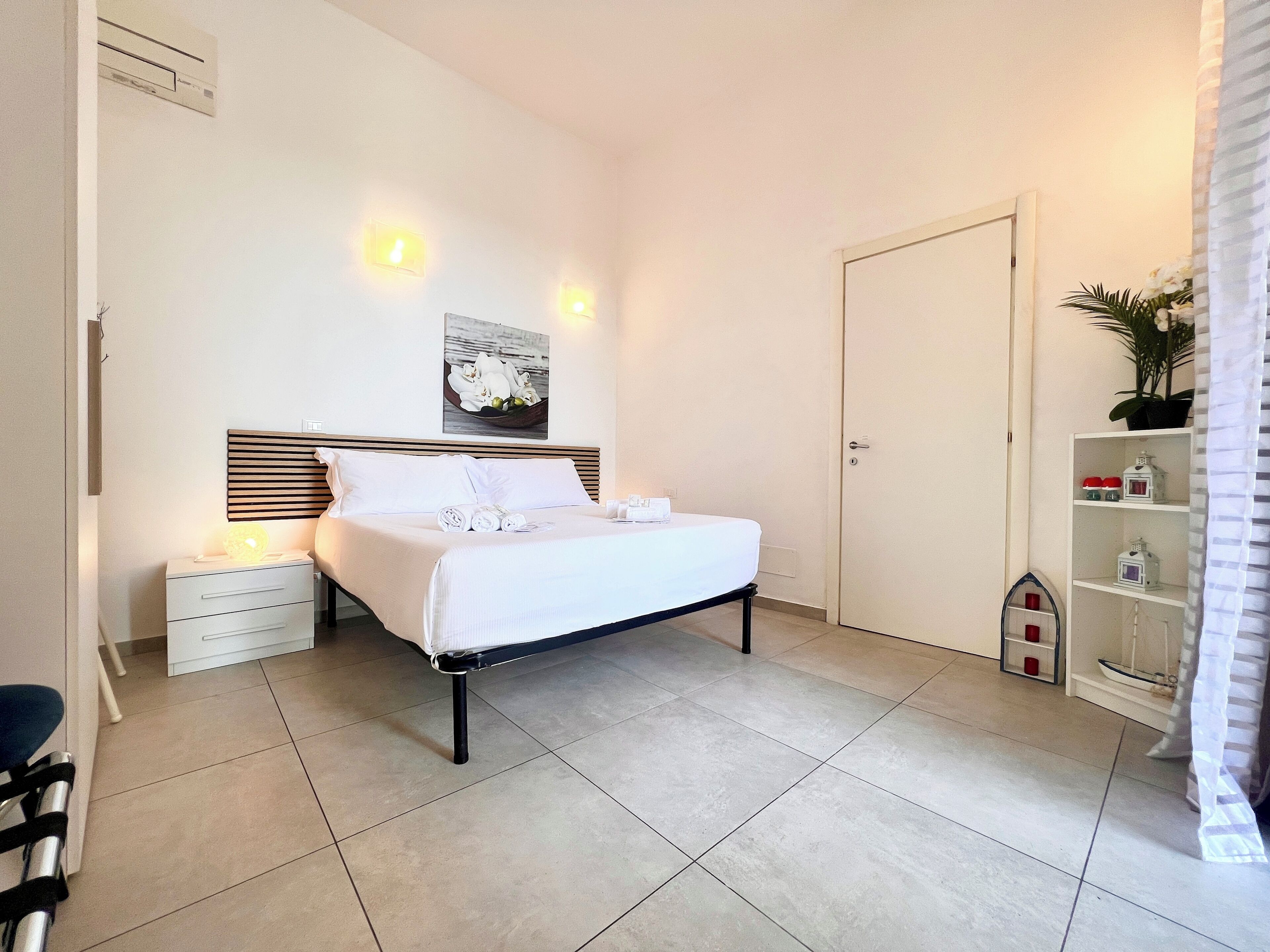 Une chambre d’hôtel bien rangée, avec un lit, des tables de chevet, une petite table d’appoint, un climatiseur fixé au mur, un tableau encadré et une petite étagère avec des objets décoratifs.