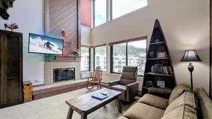 Un salon spacieux avec de hauts plafonds, une cheminée, de grandes fenêtres, un téléviseur et une bibliothèque.